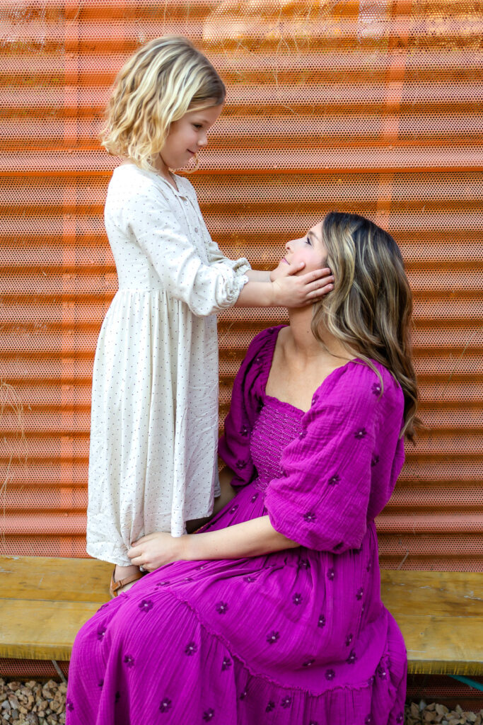 Mother cuddling daughter while standing beside warm orange wall during Palo Alto motherhood photos relaxed portrait session