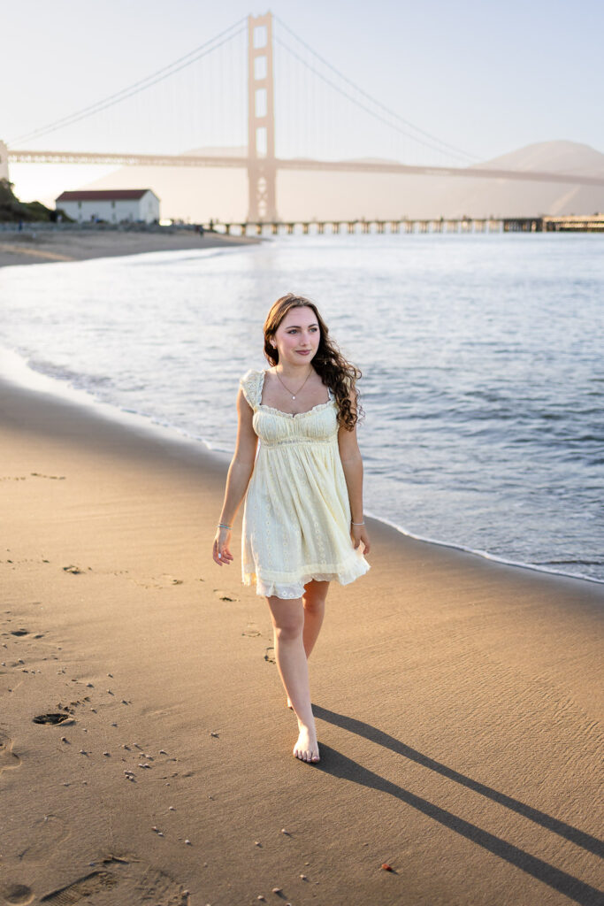 Girl walking along the shore at Crissy Field beach in a butter yellow dress with the Golden Gate Bridge behind her