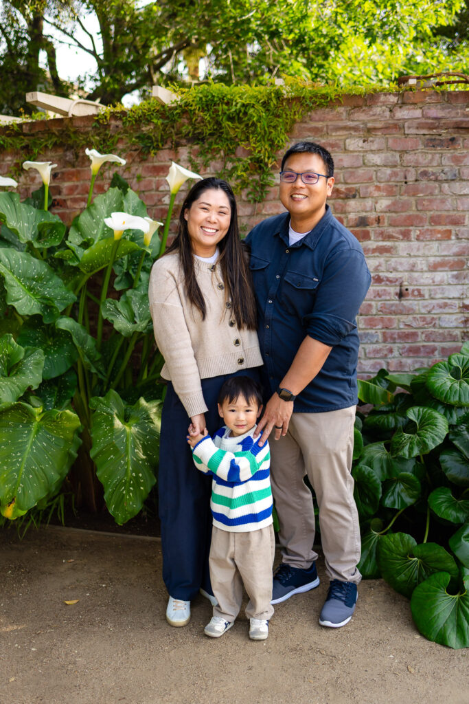 A family of three posing in front of lush greenery and a brick wall at Gamble Garden in Palo Alto.