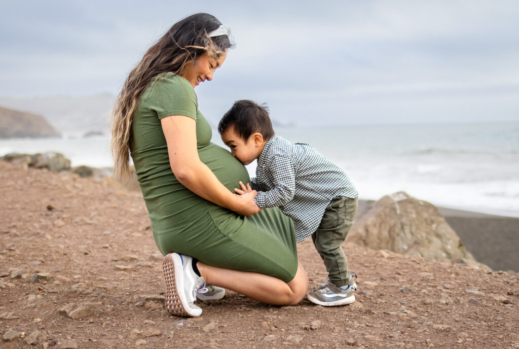 A mother in a green maternity dress kneeling to hug her young son on an outdoor nature trail.
