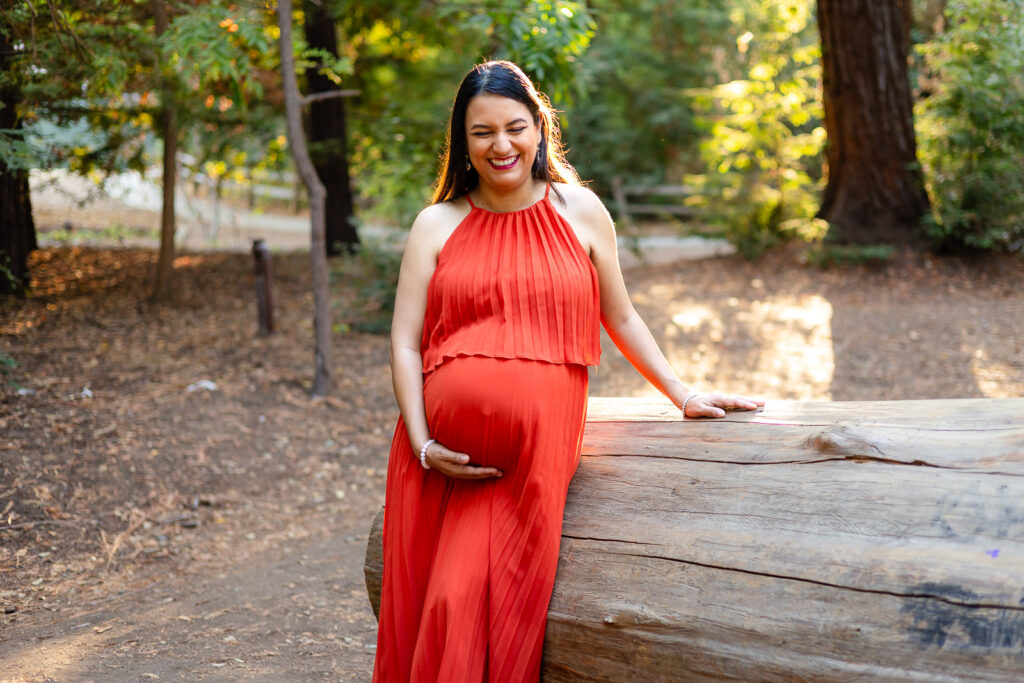 Expecting mother in a orange red dress leaning against a log under filtered sunlight on a wooded trail.