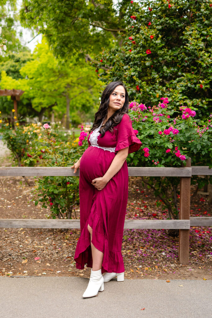 An expecting mom posing next to a rustic fence wearing a deep jewel-toned burgundy maternity dress in a park on the SF Peninsula.
