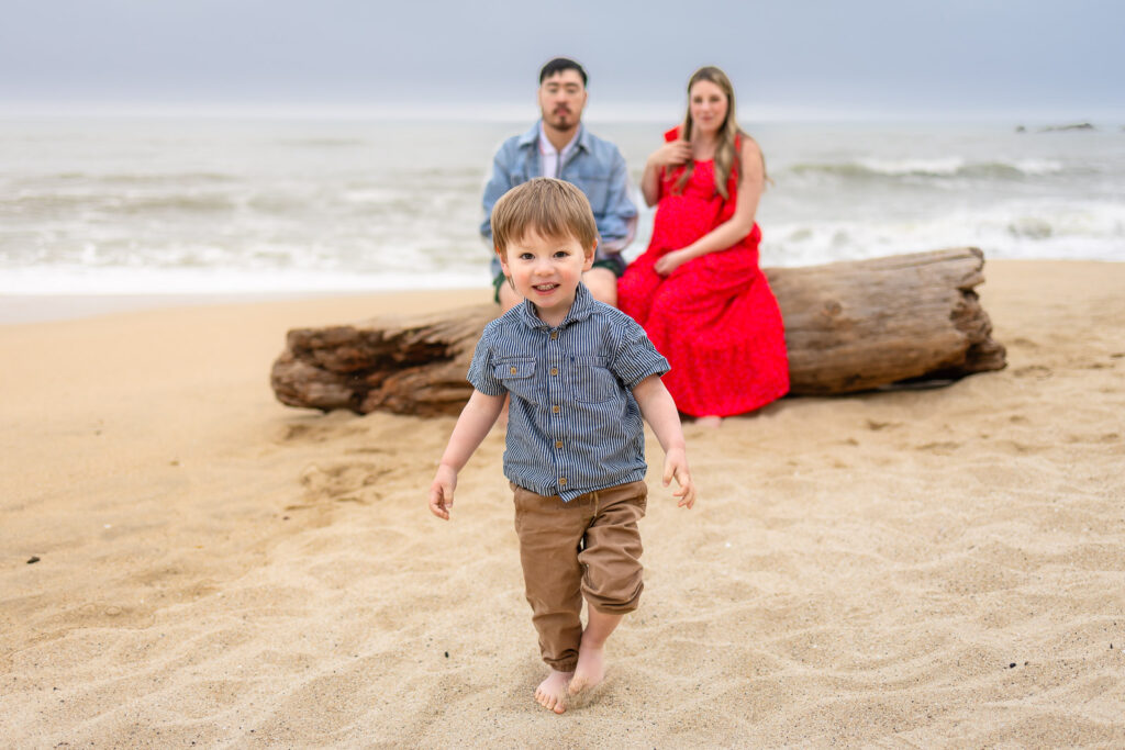 A smiling toddler runs barefoot across the sand while his parents sit together on driftwood near the ocean during a family maternity session.