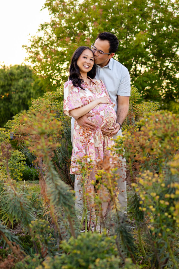 A partner holding his pregnant wife in a soft, textured cream-colored sweater and dress set during a sunset session in San Mateo County.