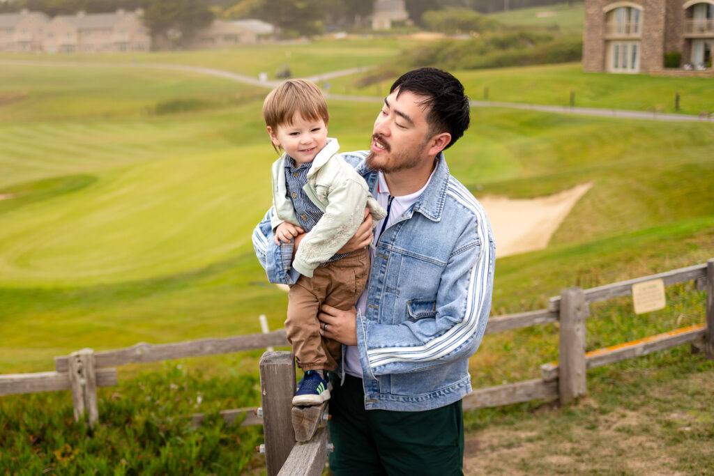 A father holds his young son while standing beside his pregnant partner near a coastal fence.
