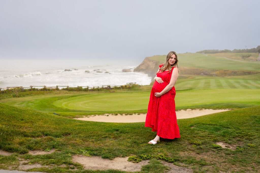 An expecting mother in a striking red dress standing on a coastal cliff overlooking the ocean in Half Moon Bay.
