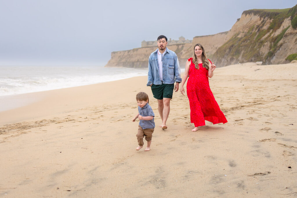 A mother, father, and toddler walking along a wide sandy beach during a coastal maternity photography session.