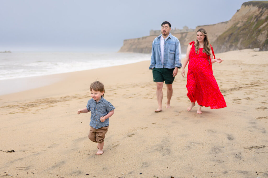 An expectant mother in a red dress walks along the sand with her partner and young son near the shoreline.