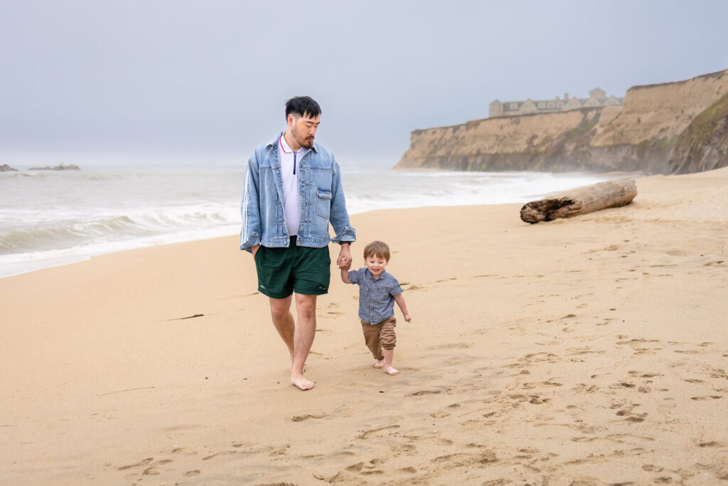 A father holds his toddler son's hand as they walk barefoot along a sandy beach with coastal cliffs in the background in Half Moon Bay.