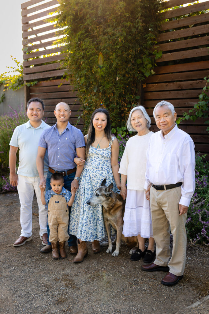 Three-generation extended family posing outdoors in San Mateo County wearing a coordinated white, khaki, and light blue color scheme with their family dog.