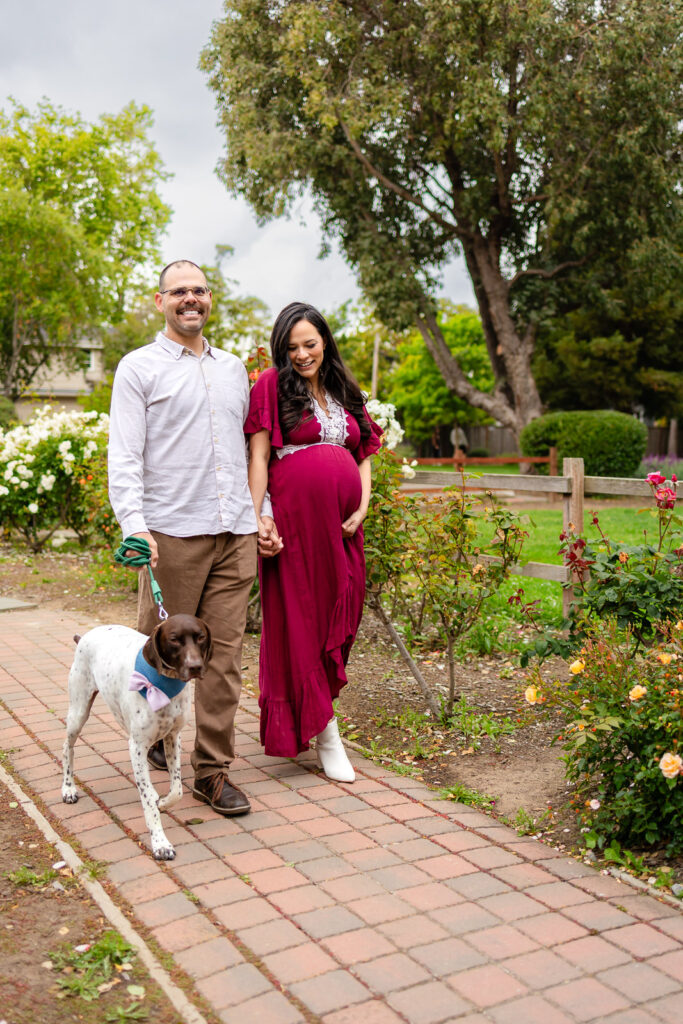 A couple walking their dog down a garden trail; the pregnant woman is in a long burgundy dress and the partner is in a neutral button-down shirt.