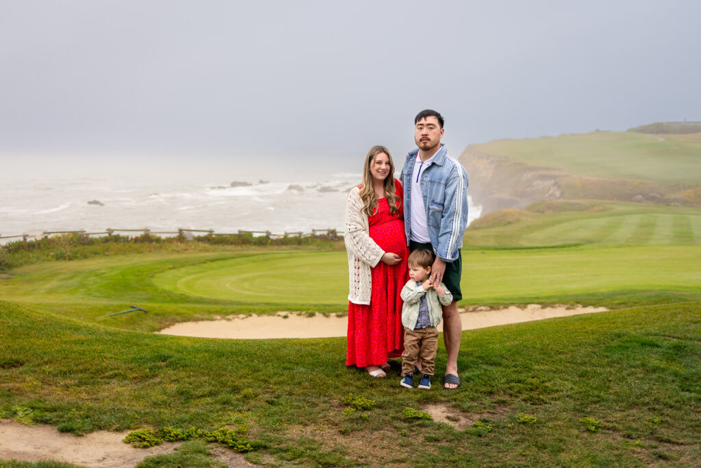 An expecting couple stands together on a coastal overlook with ocean views behind them.