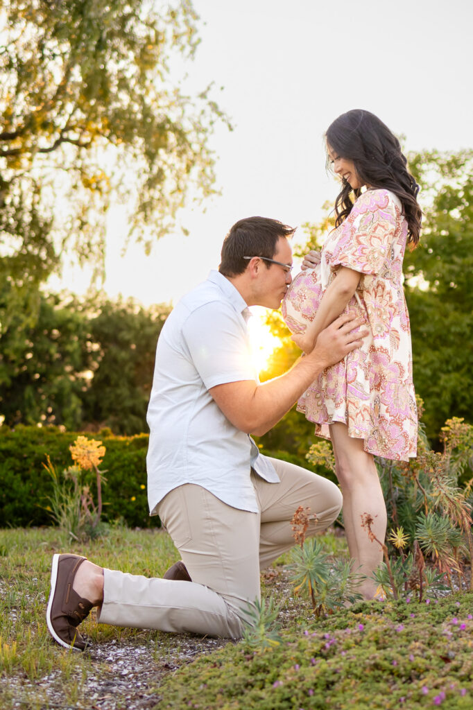 A husband kneels to kiss his wife's pregnant belly in a golden field during a sunset maternity session.