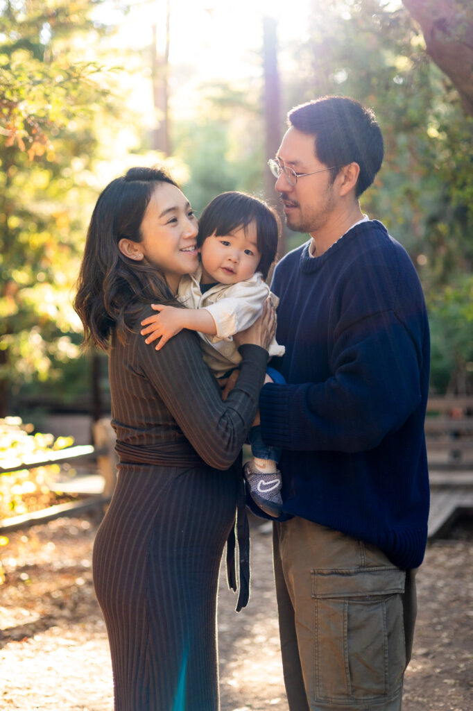 A happy family holding hands and laughing together under sunlit trees during a San Mateo County photography session.