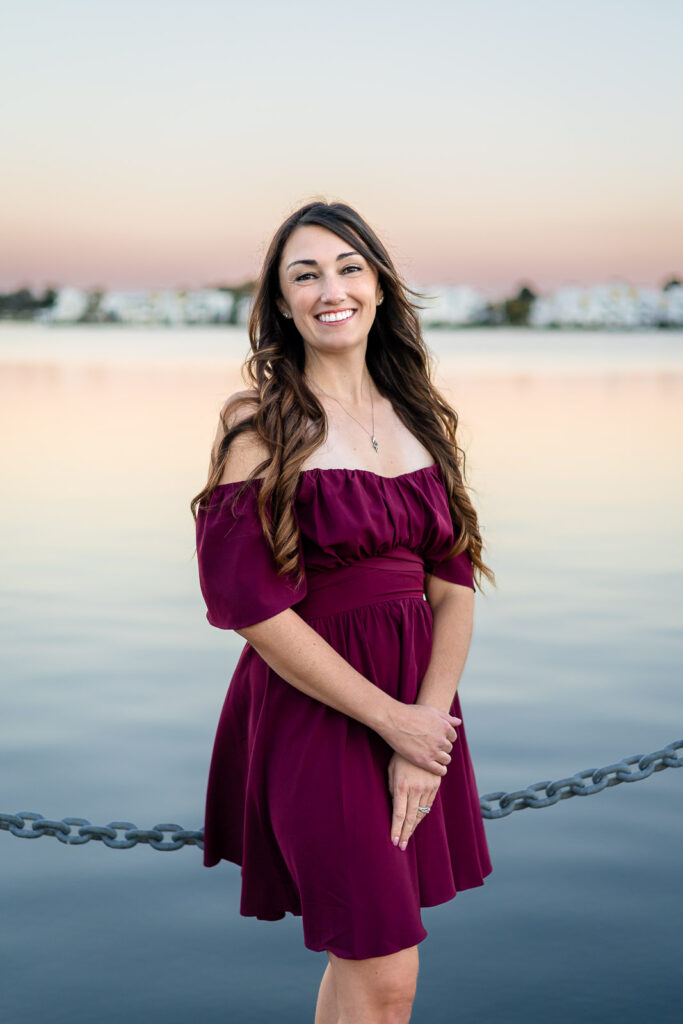 Senior girl in a burgundy dress standing by the water at sunset
