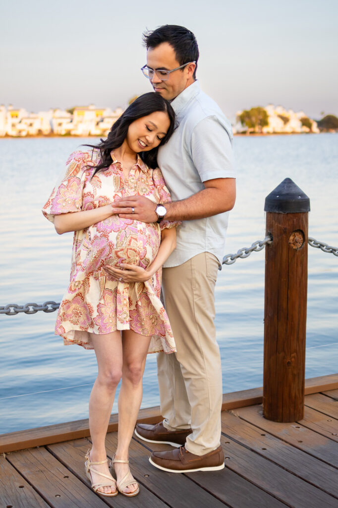 Expecting couple sharing a close embrace on a wooden boardwalk dock over the water at sunset.
