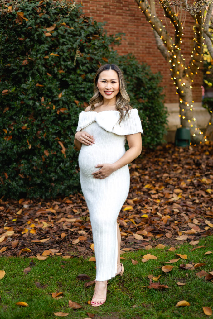 An expecting mother looking down at her bump wearing a timeless white knit maternity gown against lush greenery in Woodside
