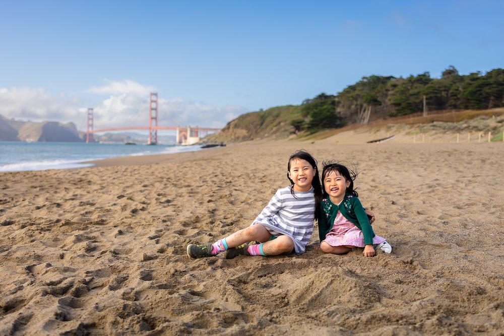 Two sisters sitting together on Baker Beach with the Golden Gate Bridge in the background on a sunny day in San Francisco.