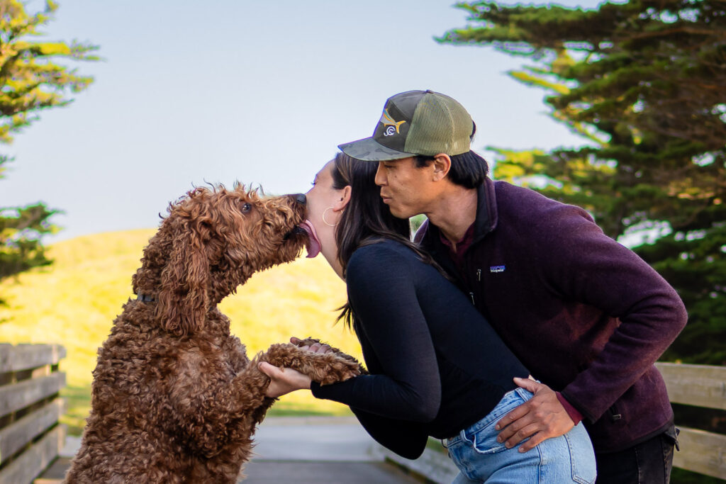 Couple sharing a kiss while their dog reaches in with an affectionate lick.