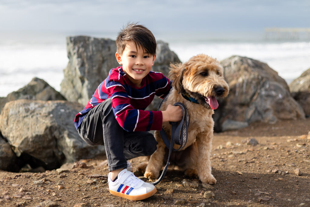 Young boy smiling beside his Goldendoodle on a rocky beach.