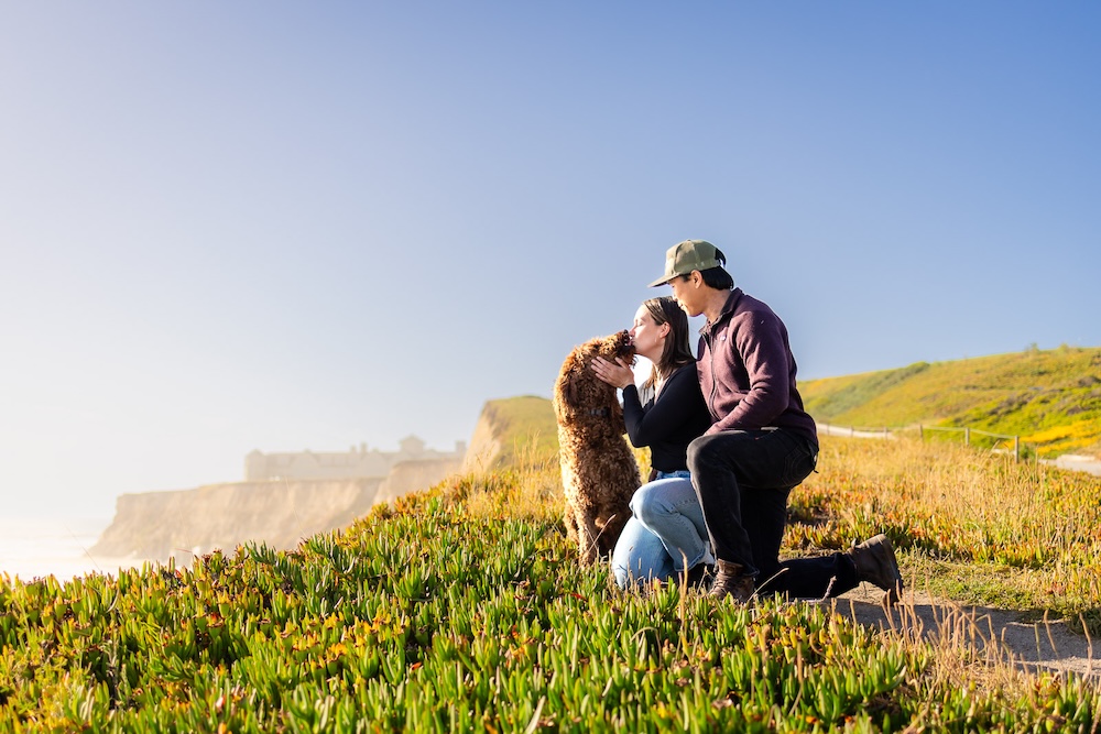 Couple kneeling with their Labradoodle overlooking the California coastline.