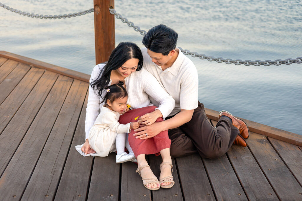 Family of three sitting on boardwalk wearing neutral white and cream and blush red