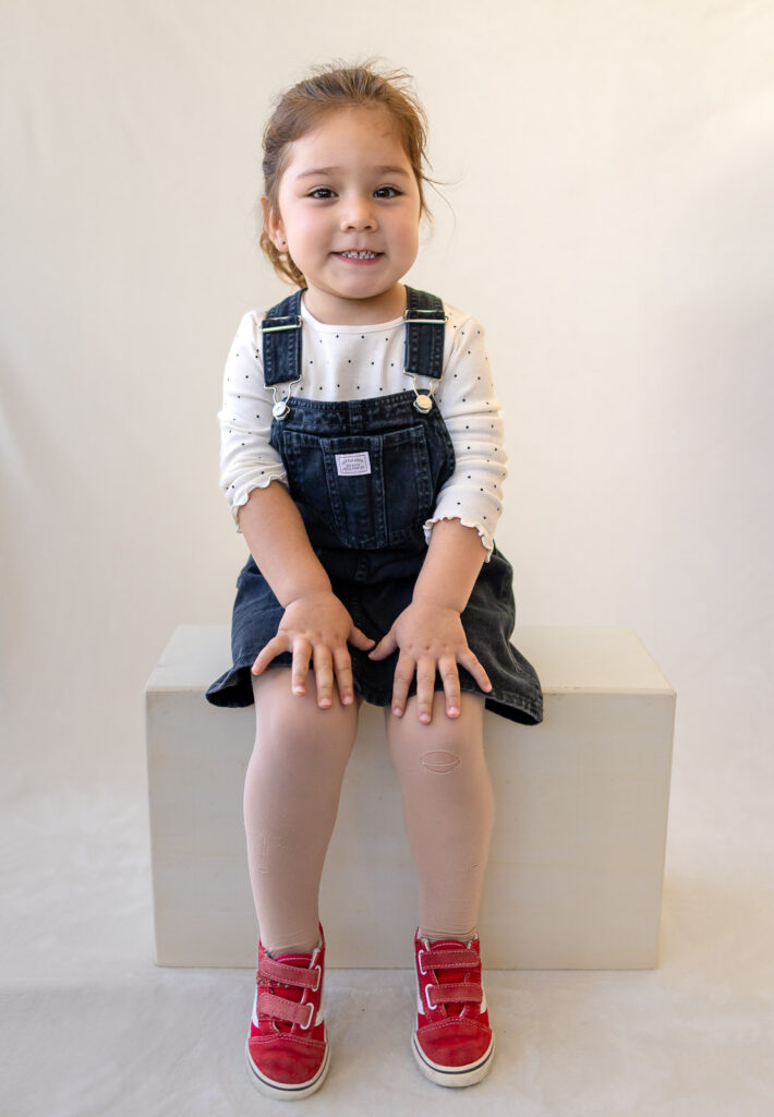 Preschool girl wearing a denim overall dress layered over a long sleeve shirt.