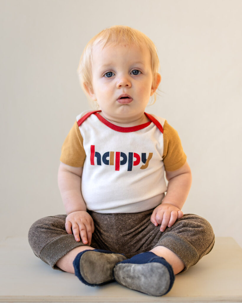 A baby boy in a shirt that reads happy sits for his portrait.