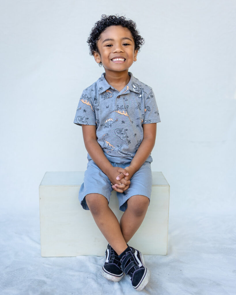 A smiling preschool boy in a dinosaur print shirt poses for his school portrait with a Bay Area preschool photographer.