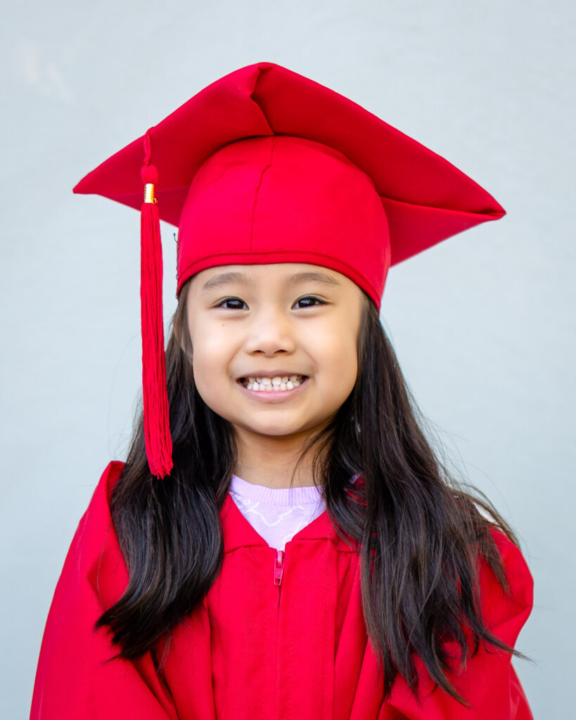 A preschool girl smiles in her red cap and gown for preschool graduation portraits.