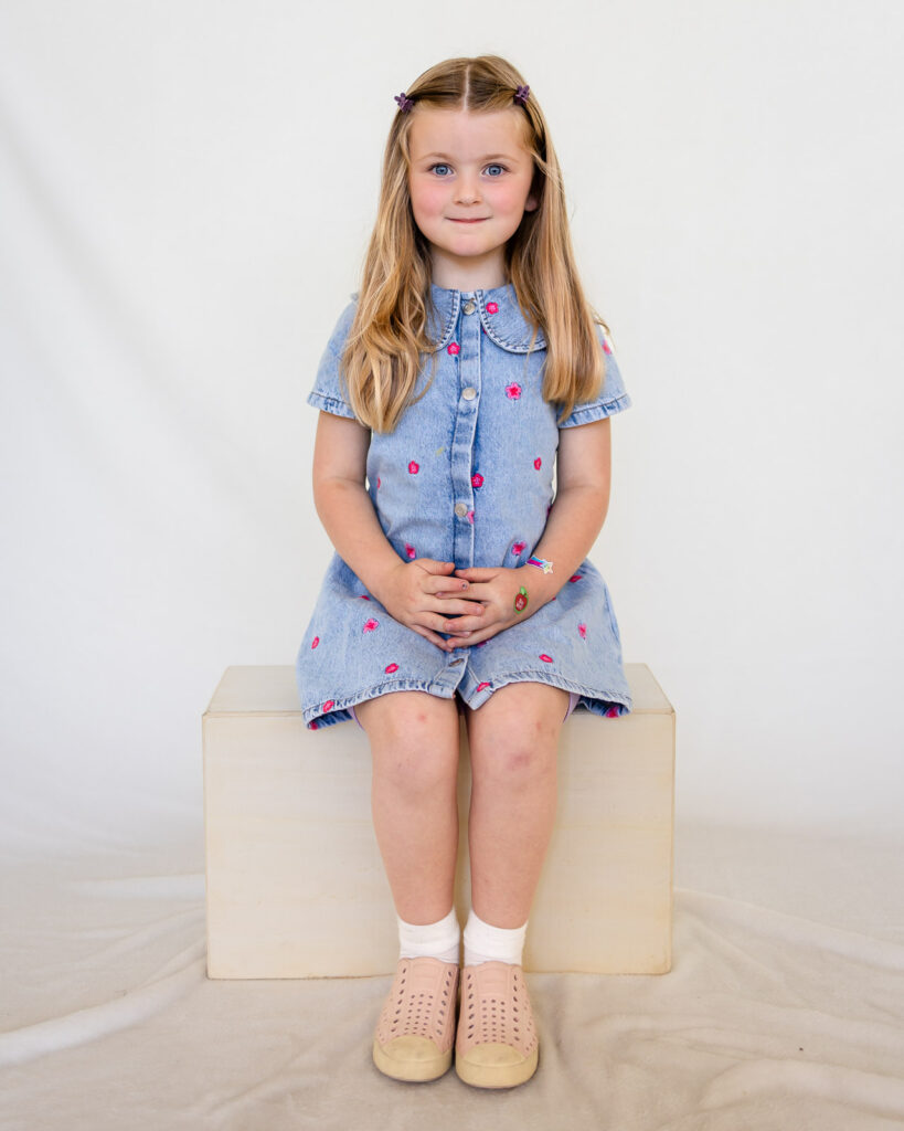 A preschool girl in a denim floral dress poses for her school portrait with a San Francisco school photographer.