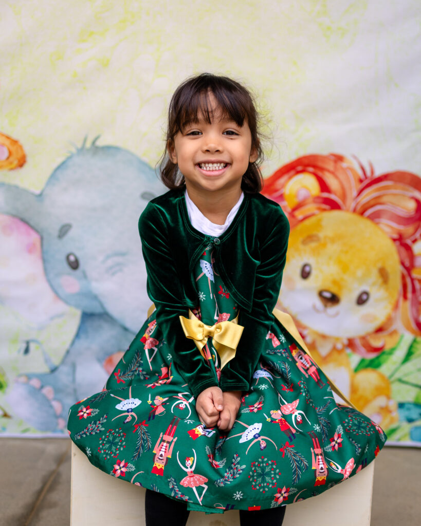 A preschool girl twirls in a green velvet holiday dress in front of a playful watercolor animal backdrop for a Bay Area school photographer.