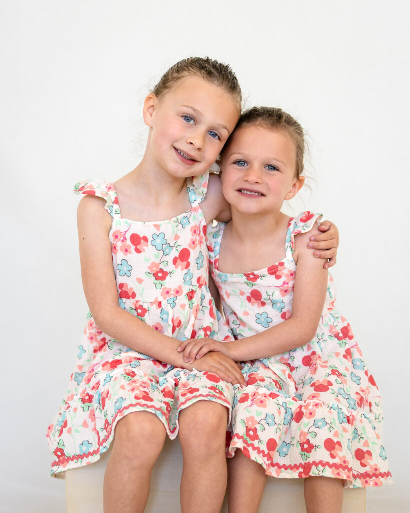 Two sisters in matching floral dresses hug for a sibling portrait with a Bay Area school photographer.