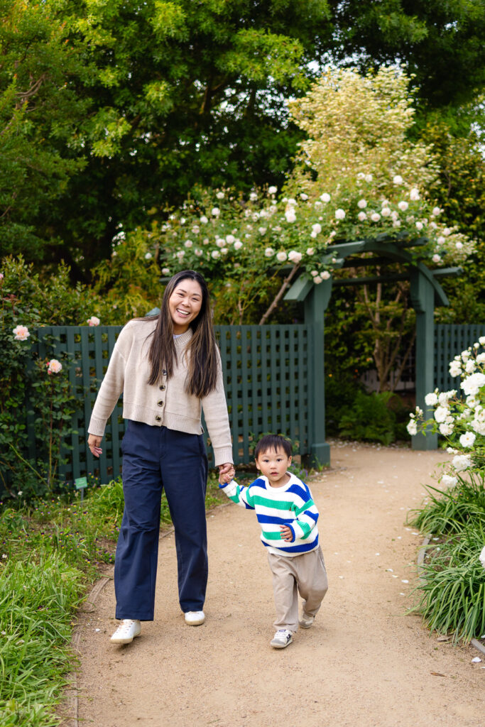 A mother holds her toddler's hand as they walk together near a rose covered arbor, guided by a Palo Alto family photographer at Gamble Garden.