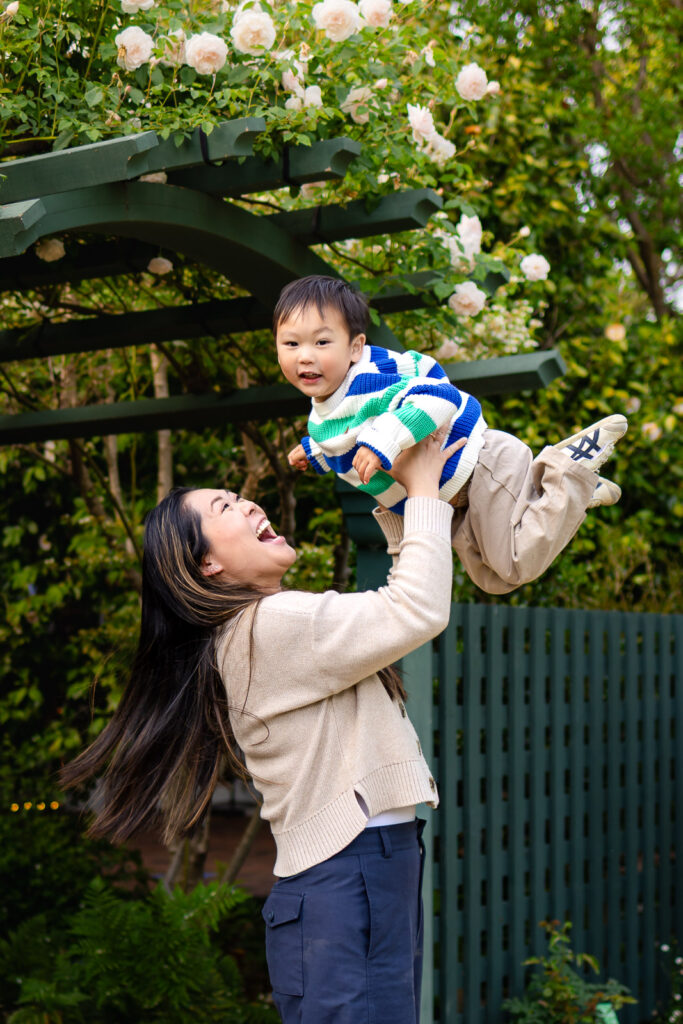 A mother laughs while lifting her toddler into the air under a blooming rose arbor during a Gamble Garden Palo Alto family photos session.