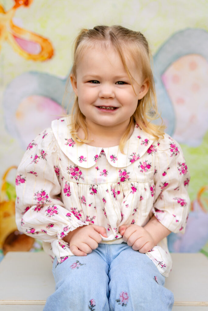 A toddler girl in a floral blouse smiles for her preschool portrait with a San Francisco preschool photographer.