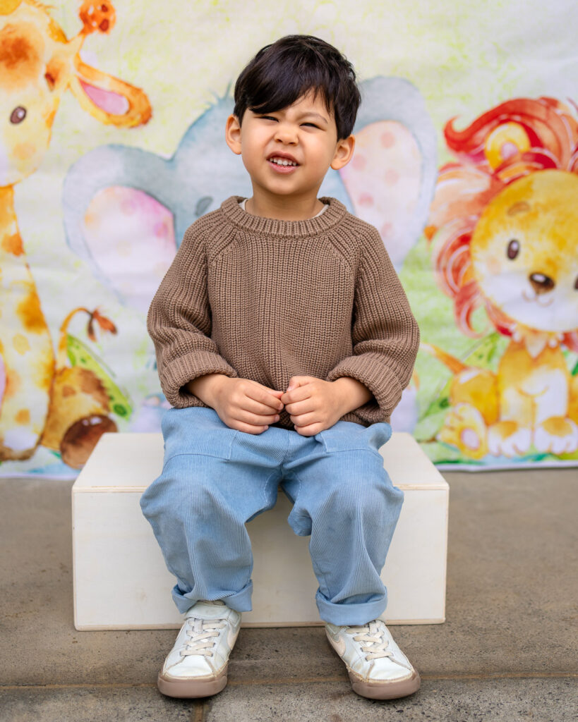 A laughing preschool boy in a brown sweater poses in front of a watercolor animal backdrop.
